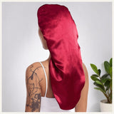 Une femme vue de dos portant un bonnet long en satin rouge éclatant, dans un décor intérieur clair avec une plante verte
