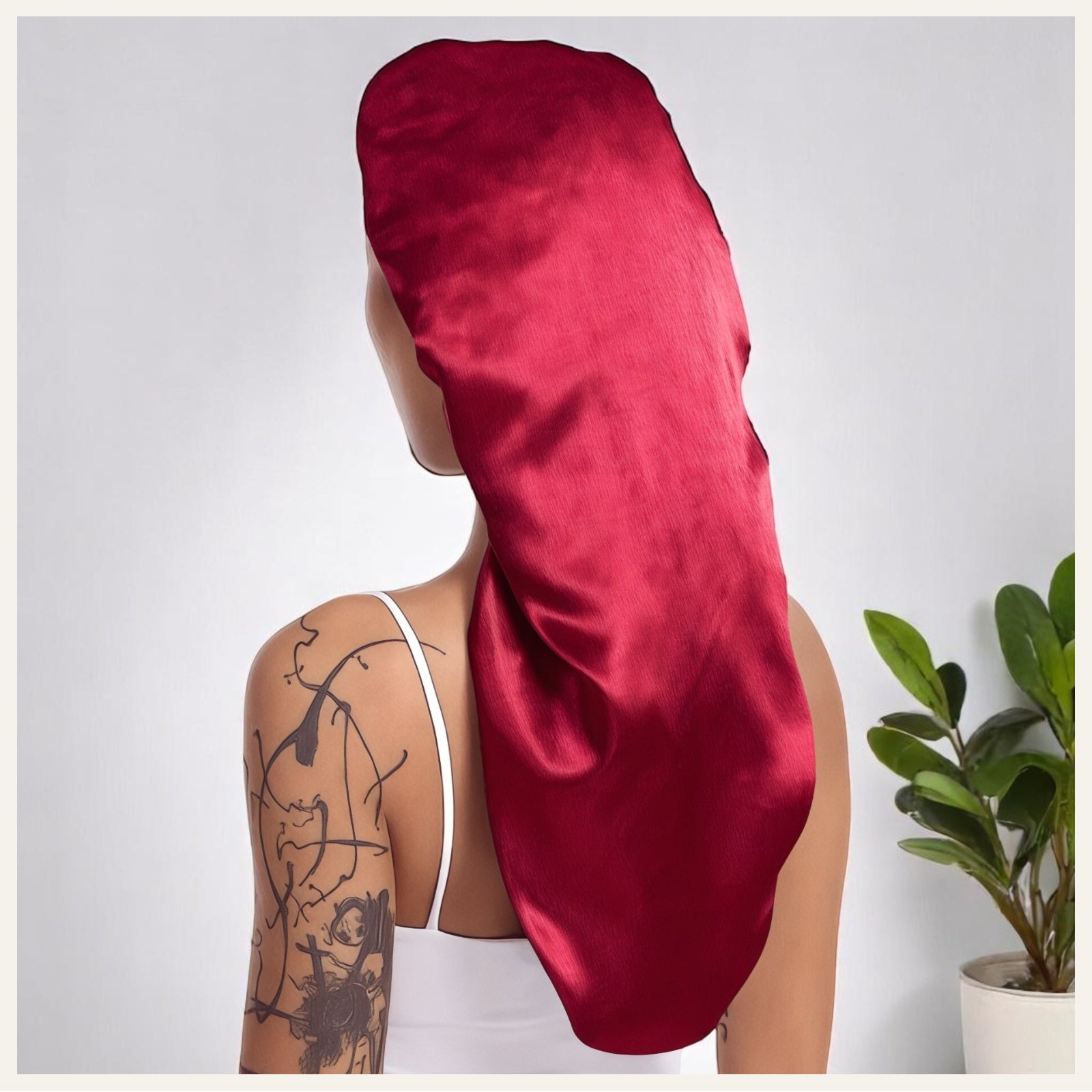 Une femme vue de dos portant un bonnet long en satin rouge éclatant, dans un décor intérieur clair avec une plante verte