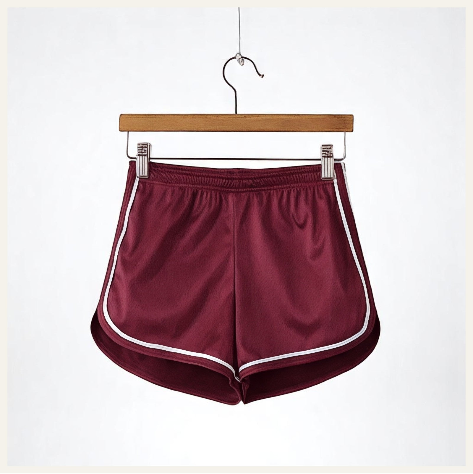 Un short de pyjama en satin rouge bordeaux avec liserés blancs, suspendu sur un cintre en bois contre un fond blanc