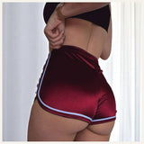 Une femme portant un pyjama short en satin rouge bordeaux à liserés blancs, photographiée de dos dans un intérieur lumineux