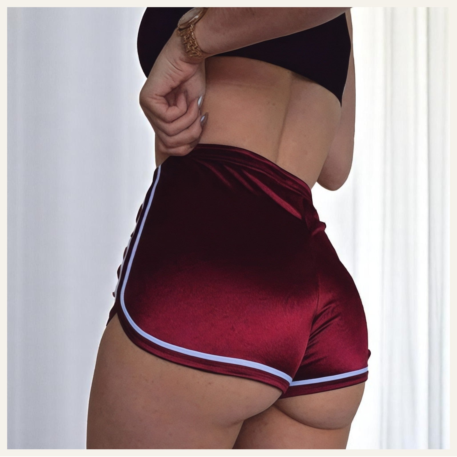 Une femme portant un pyjama short en satin rouge bordeaux à liserés blancs, photographiée de dos dans un intérieur lumineux